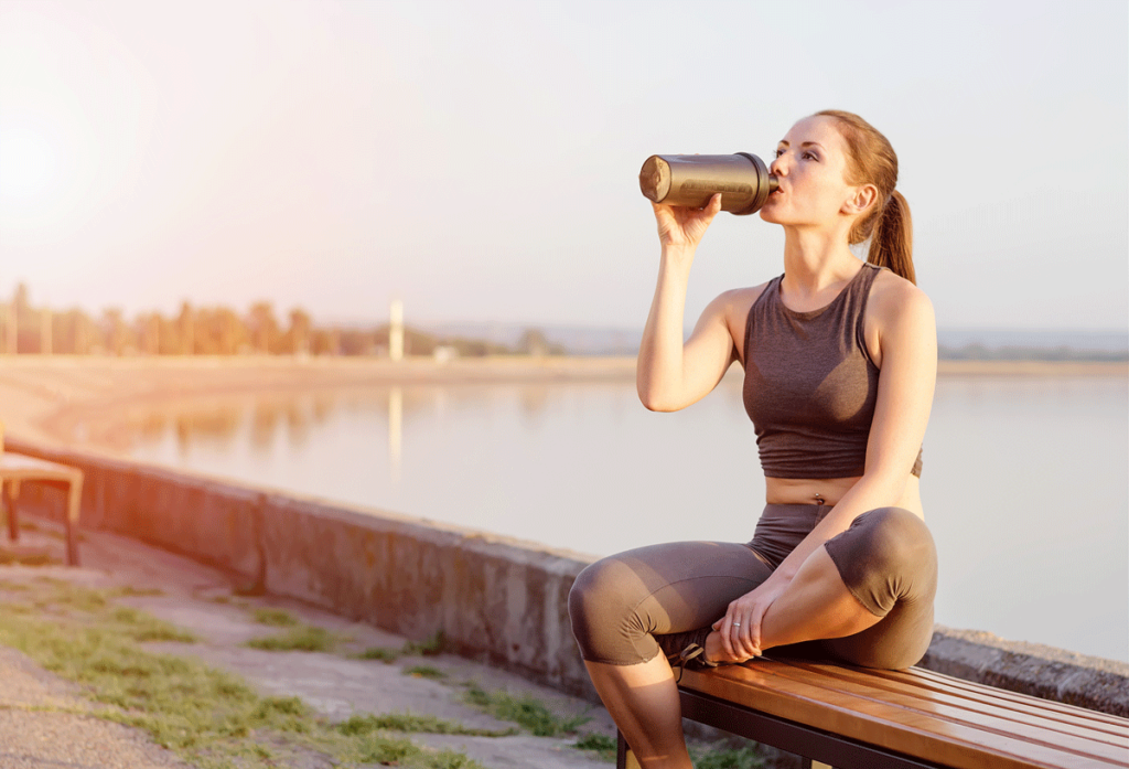 Junge Sportlerin sitzt nach dem Training auf einer Bank und trinkt Proteinshake