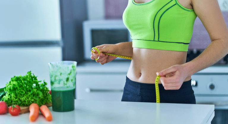 Junge Frau hält in der Küche ein Maßband um ihre Taille, vor Ihre ein grüner Smoothie und Gemüse