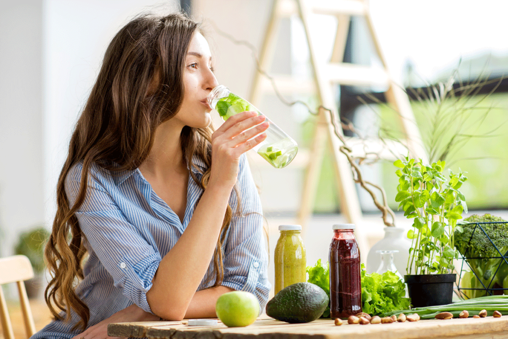 Junge Frau trinkt einen Wasser mit Limetten und Zitronen, vor ihr verschiedenen Smoothies